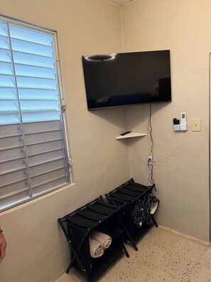 A room with a TV mounted on the wall, a window with blinds, a black shelf unit with rolled-up towels, and a power outlet with a phone charger. - Private 7-Acre El Yunque Retreat | Generator (luquillo)