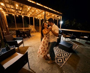 Un patio exterior con muebles de madera, luces colgantes y un tapete estampado.
