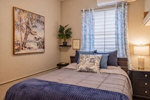 A bedroom with a bed, a nightstand, a lamp, a window with curtains, a plant, and a painting on the wall. - Private 7-Acre El Yunque Retreat | Generator (luquillo)