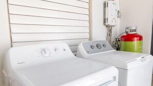Una lavadora y secadora Hotpoint blancas de carga frontal, con perillas de control, un extintor de incendios rojo y un enchufe eléctrico blanco sobre una pared blanca.