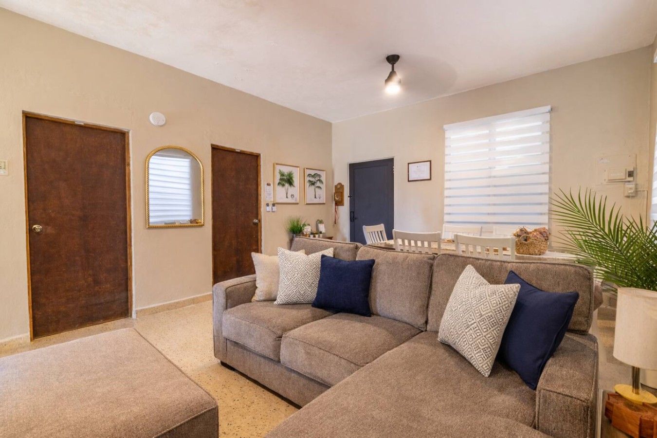 A living room with a sectional sofa, a dining area with a table and chairs, and a large window with blinds.
