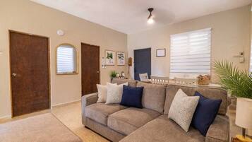 A living room with a sectional sofa, a dining area with a table and chairs, and a large window with blinds.