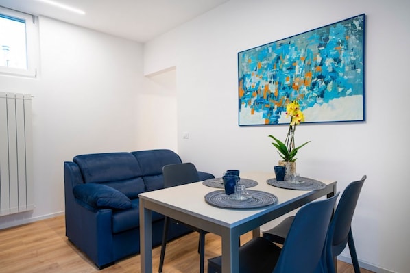 A modern living room with a blue sofa, a dining table with blue chairs, a painting on the wall, and a potted plant.