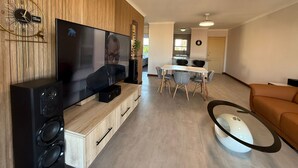 Egy modern nappali, melynek berendezéséhez tartozik egy síkképernyős televízió, fa borítású fal, étkező, konyha és egy egyedi, fehér kávézóasztal.