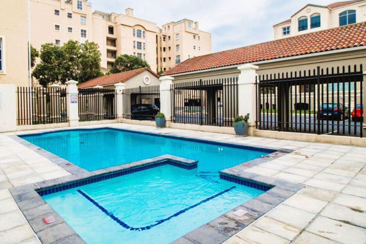Une piscine rectangulaire bordée de carreaux, entourée d’une allée pavée. On aperçoit plusieurs immeubles d’appartements en arrière-plan, avec des balcons et un toit recouvert de tuiles rouges.