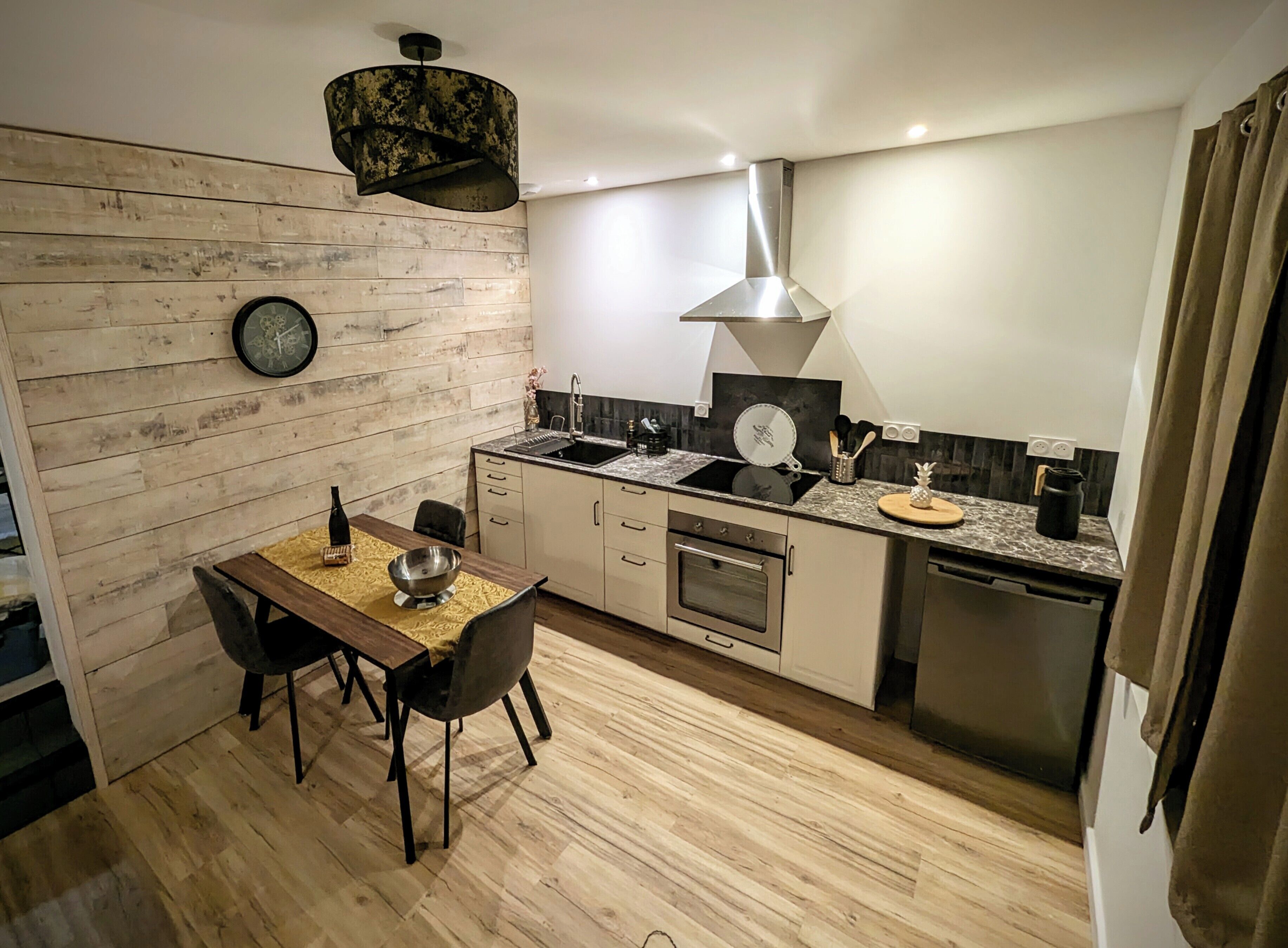 Een compacte keuken met een houten wand, een eettafel met stoelen, een spoelbak, een fornuis en een vaatwasser.