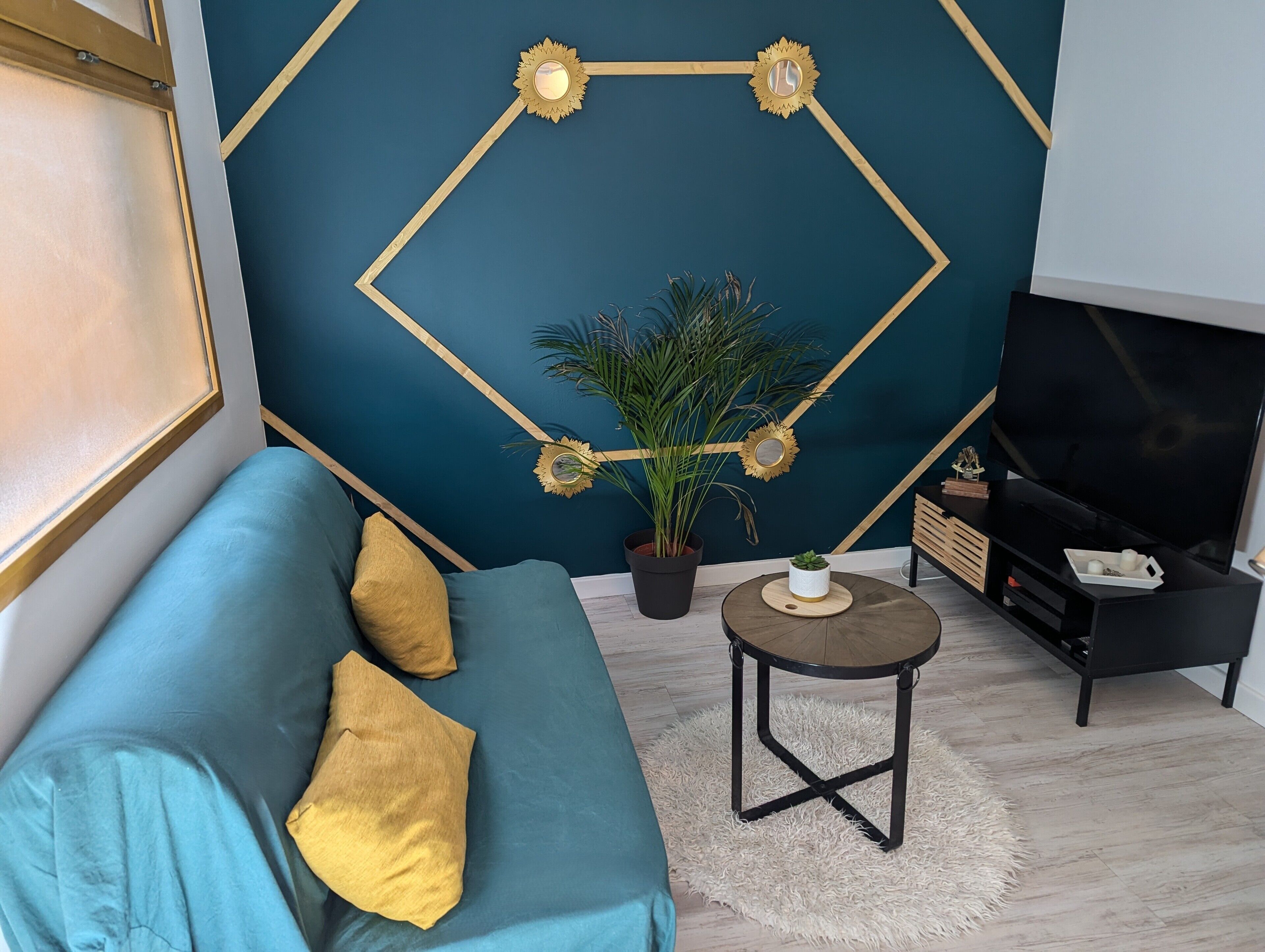 A modern living room with a teal sofa, a small round table, a potted plant, and a TV stand.