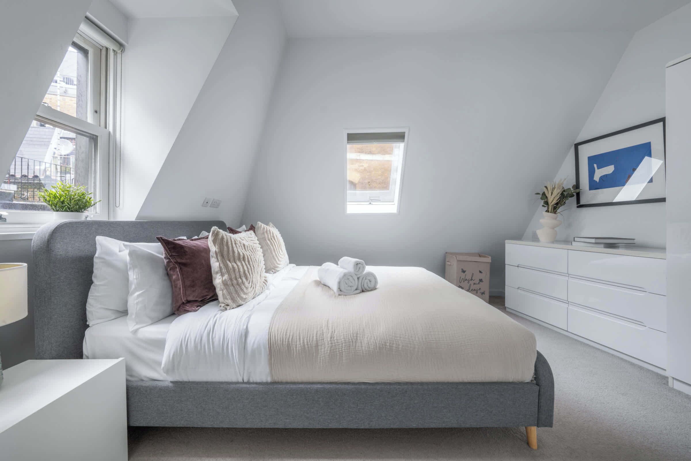 Ein modernes Schlafzimmer mit Bett, Nachttischen, Kommode, Fenster und einem gerahmten Bild an der Wand.