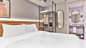 Une chambre d’hôtel avec un grand lit, une table de chevet avec une lampe, une armoire, un miroir et une salle de bain équipée d’un lavabo et d’un porte-serviettes.