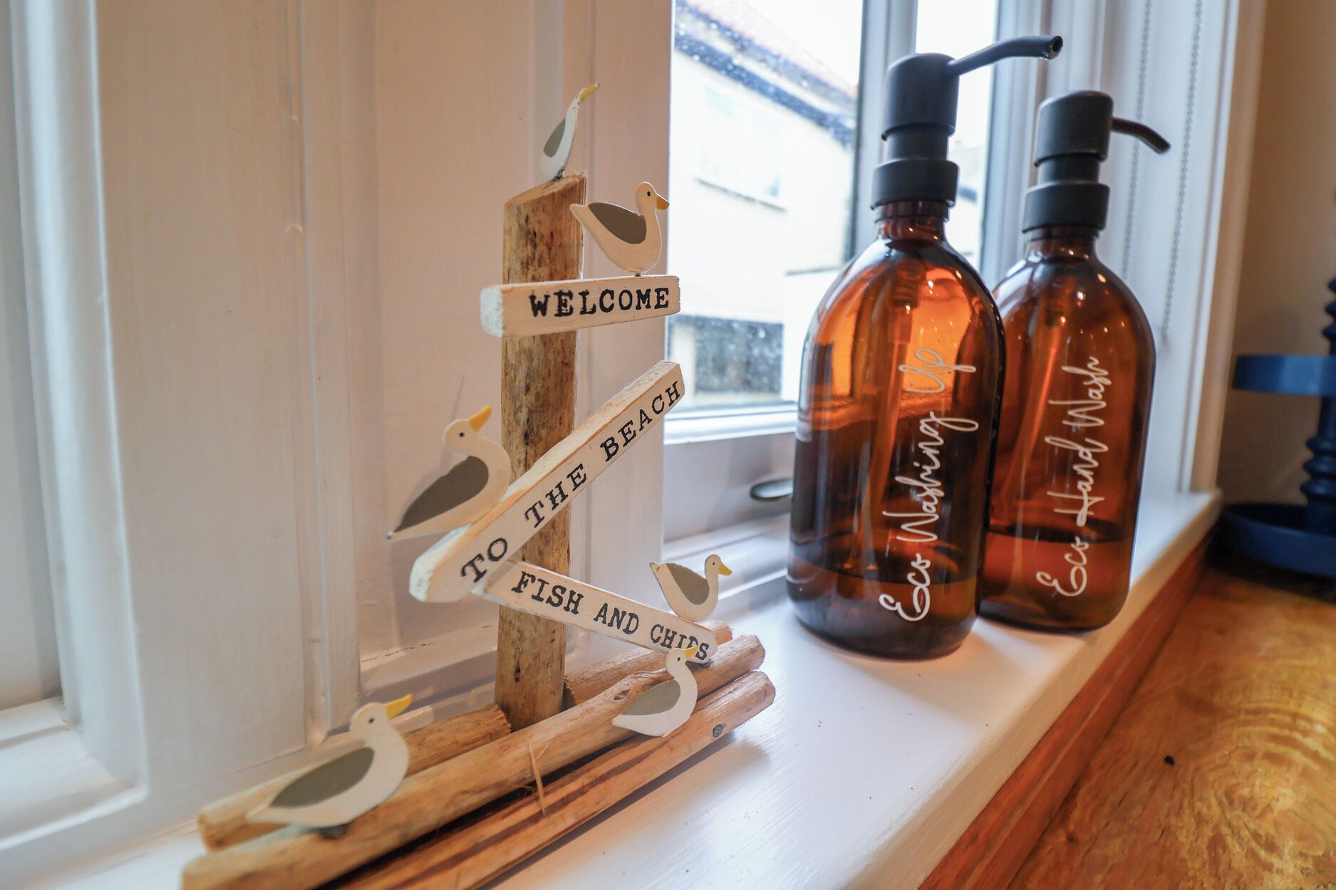 Un letrero de madera con adornos de pájaros y el texto que dice "WELCOME TO THE BEACH" y "FISH AND CHIPS". Dos botellas ámbar con dosificadores están sobre una estantería.