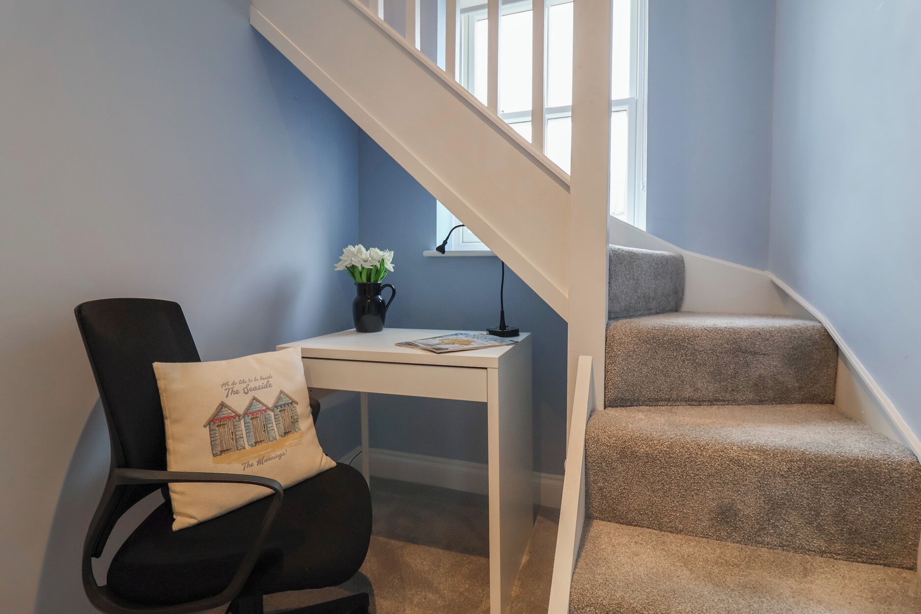 Une pièce comprenant un fauteuil de bureau noir, un petit bureau blanc, un vase avec des fleurs blanches et un escalier recouvert d’une moquette beige.