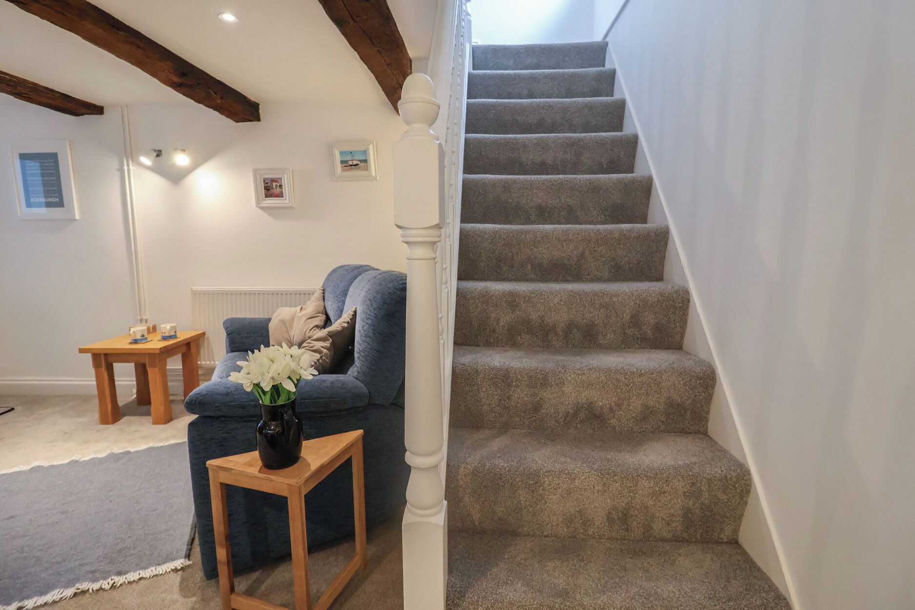 Una escalera con peldaños alfombrados, un sofá azul, una pequeña mesa de madera con un jarrón de flores y una pared blanca con cuadros enmarcados.