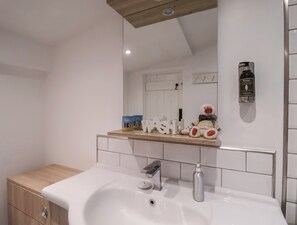 Un baño moderno con un lavamanos blanco, un mueble de tocador de madera, un espejo y un estante con objetos decorativos.