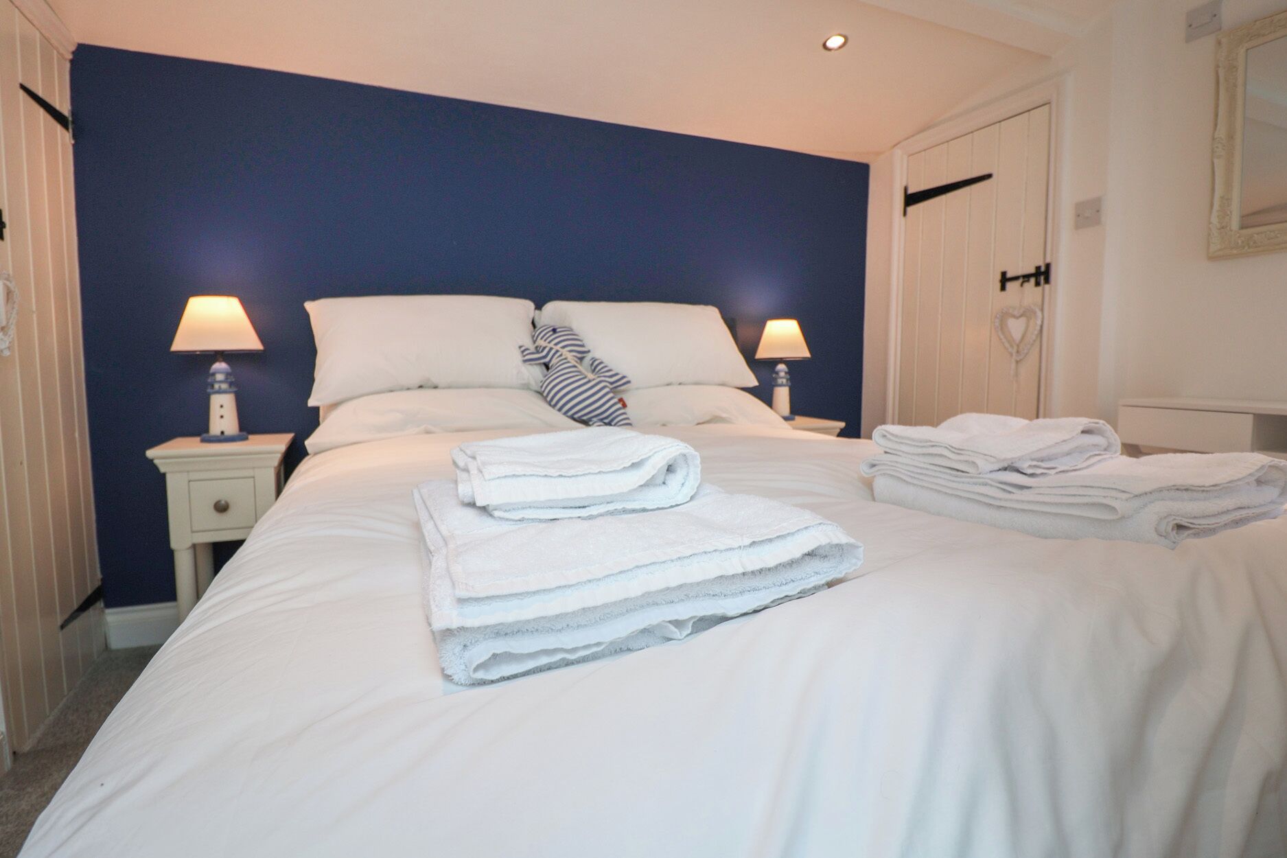 Un lit bien fait, avec du linge de lit blanc et des serviettes pliées, deux lampes de chevet et une décoration en forme de cœur accrochée au mur.