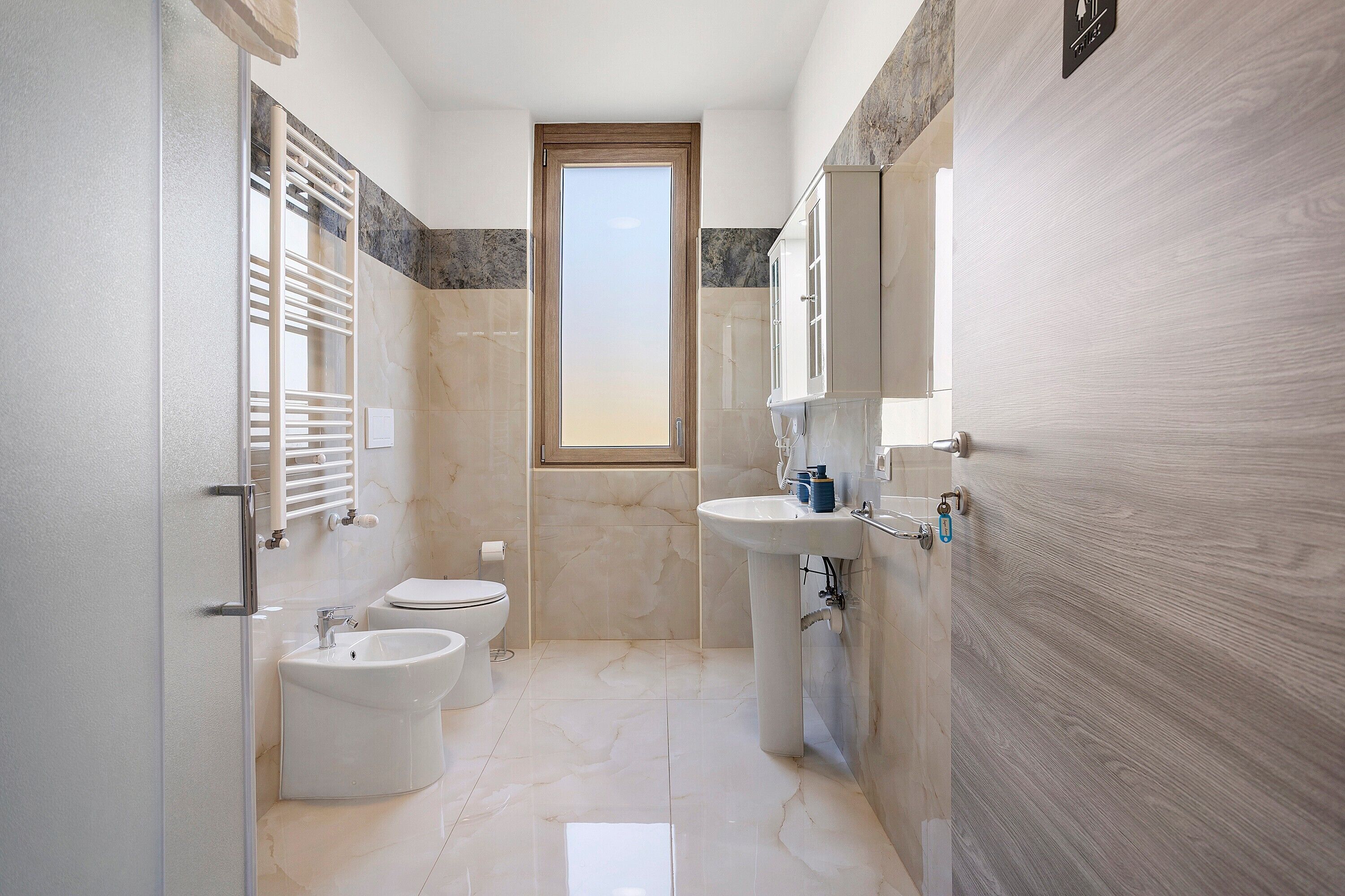 A modern bathroom with a bidet, toilet, and sink, featuring a marble floor and a wooden door.