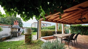 Un patio cubierto con una mesa y sillas, rodeado de follaje y una estructura de piedra.
