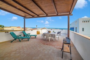 A covered outdoor seating area with hexagonal tiles, green lounge chairs, a table with chairs, and a grill. - Sunny Sea View (Melendugno)