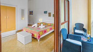 Un dormitorio con una cama, un aparador blanco, una silla de mimbre y una mesita con una botella y vasos.