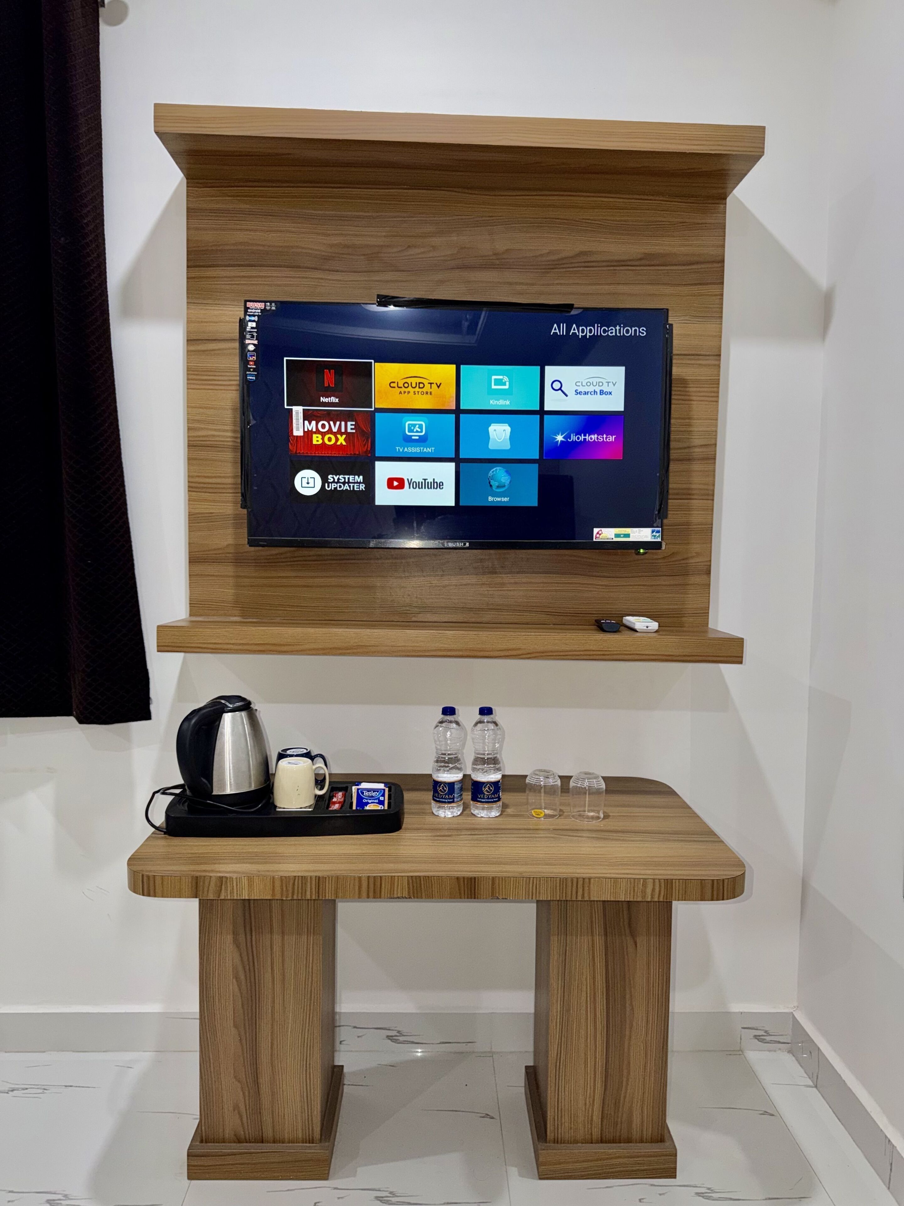 A flat-screen TV mounted on a wooden wall above a wooden table with a kettle, two water bottles, and glasses.