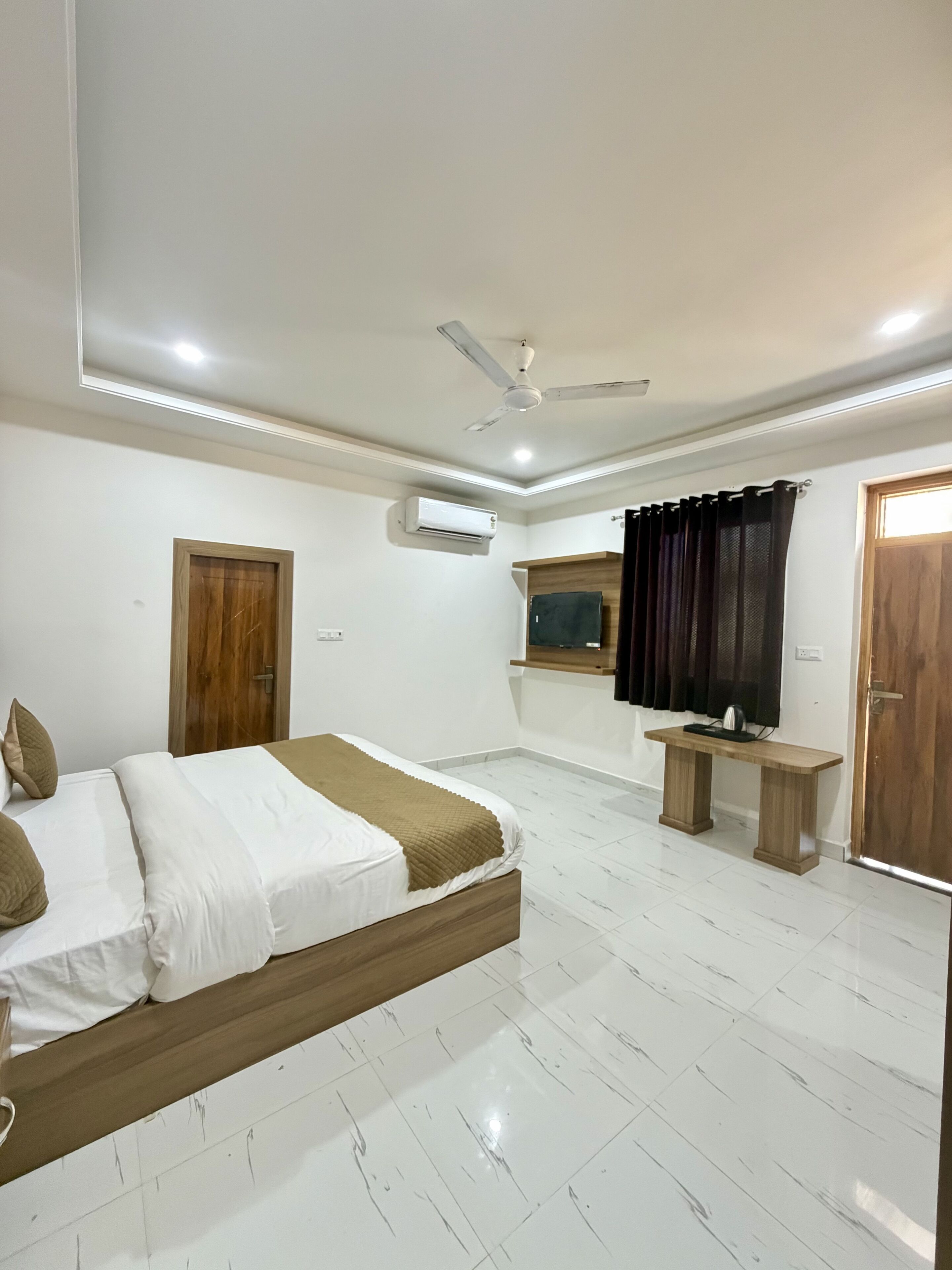 Une chambre à coucher comprenant un lit, un ventilateur de plafond, un appareil de climatisation, une porte en bois, une fenêtre avec des rideaux et une petite table en bois.