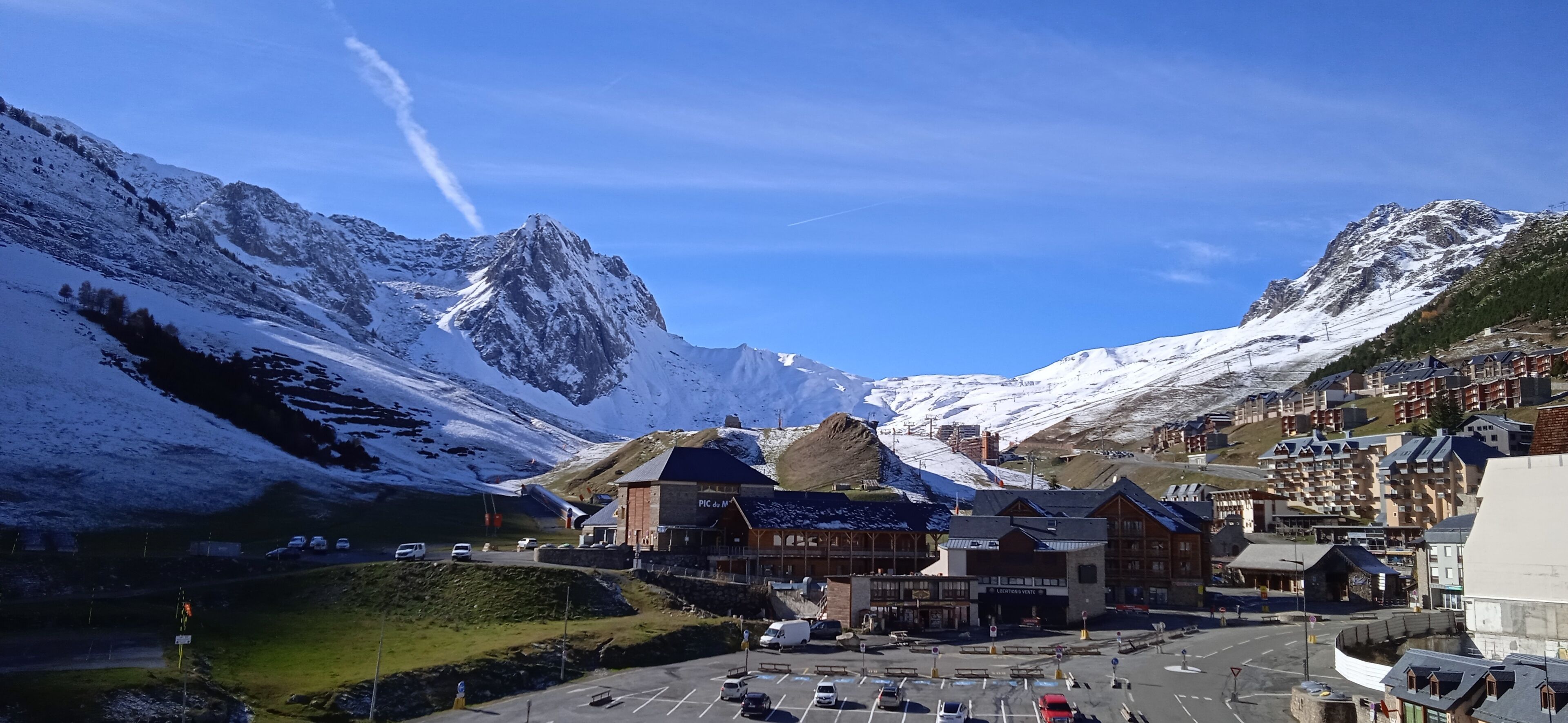 A mountain town with snow-covered peaks, a prominent hotel with a sign reading "PIC de la Montagne," and a parking lot with several cars.