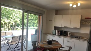 A modern kitchen with white cabinets, a dining area with a table and chairs, and a view of greenery through the sliding glass door. - Appartement 48m² 1 chambre, garage, proche du lac (Aix-les-Bains)