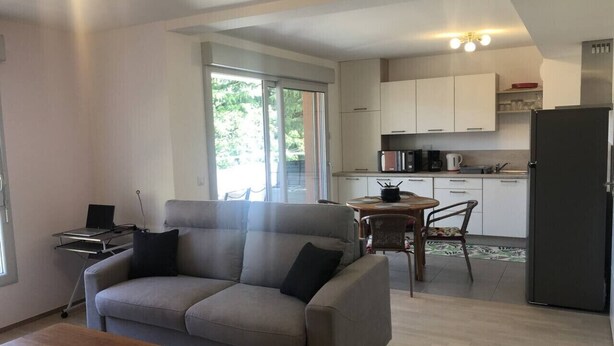 A modern living room with a sofa, a dining table, and a kitchen in the background. - Appartement 48m² 1 chambre, garage, proche du lac (Aix-les-Bains)