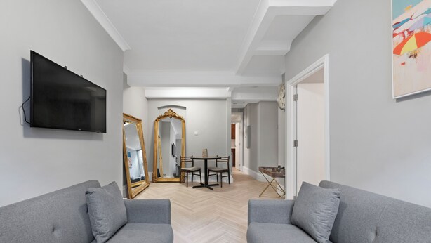 Un salon moderne avec deux canapés gris, un téléviseur à écran plat, une table à manger avec des chaises, un miroir et un parquet à motifs en chevrons.