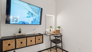 Una habitación de hotel moderna con una televisión de pantalla plana grande montada en la pared, una mesita de noche con espejo, una silla y cestas de mimbre sobre una consola negra.