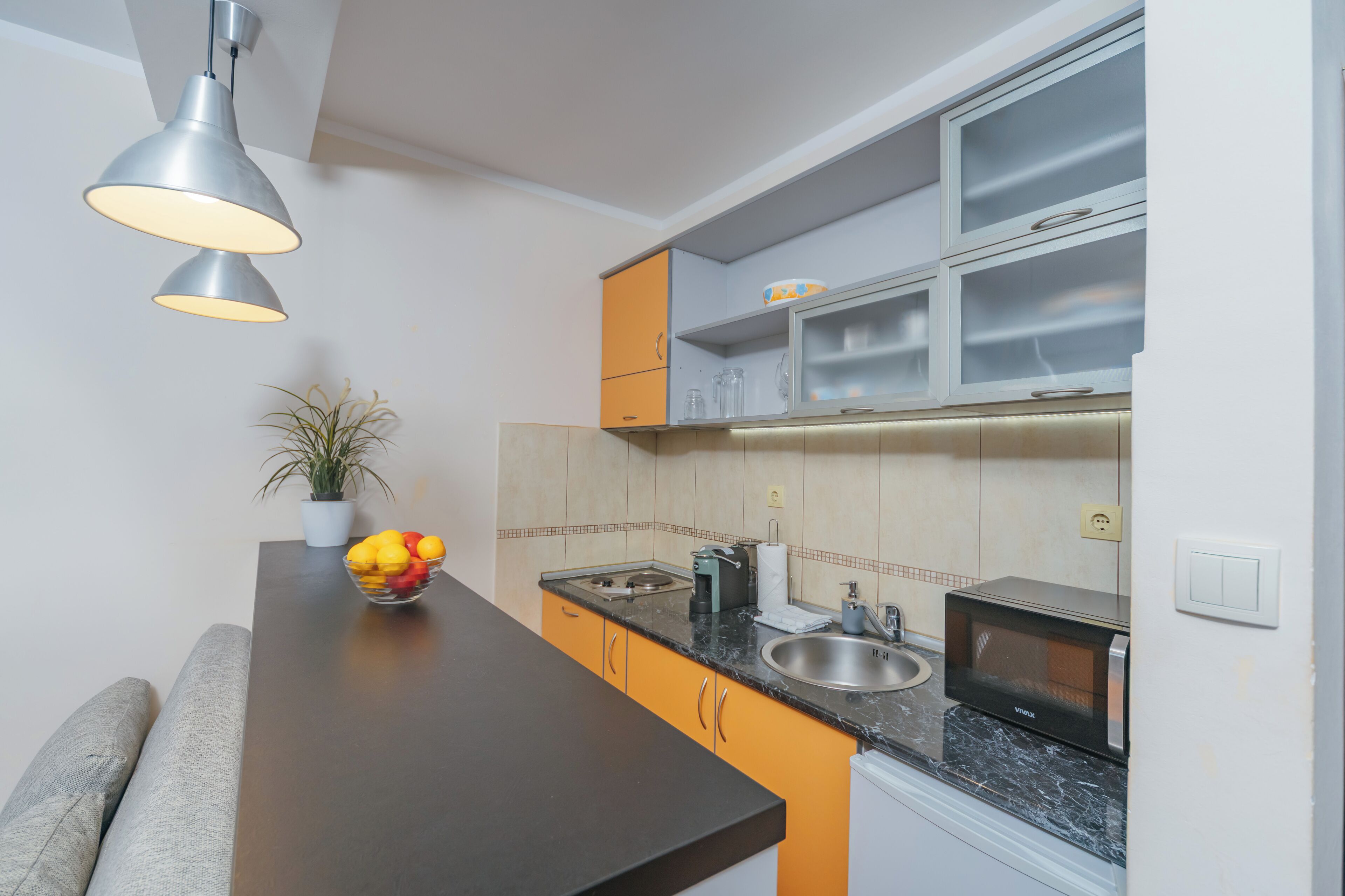 A modern kitchen with a marble countertop, a built-in microwave, and a sink. There is a bowl of fruit on the counter and a potted plant.