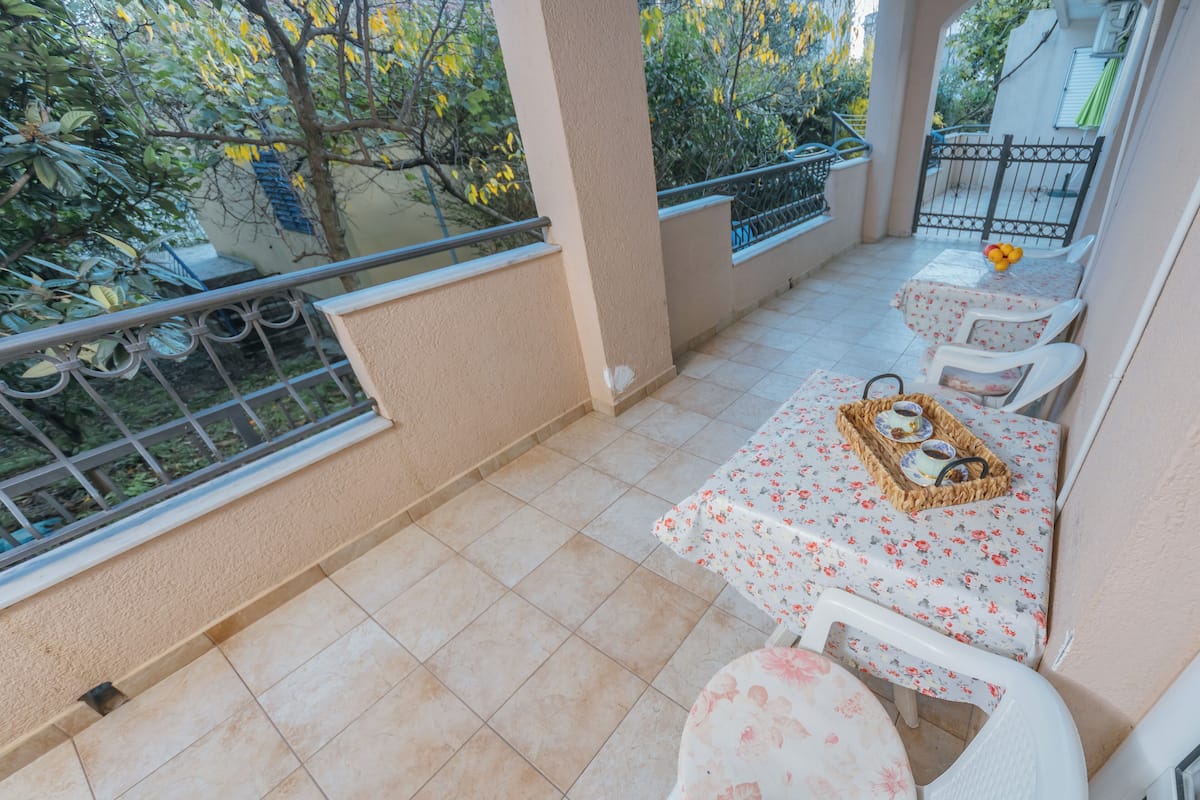 Een balkon met een betegelde vloer, twee witte stoelen, een kleine tafel met een tafelkleed met bloemenmotief, en een dienblad met bekers en een kom.