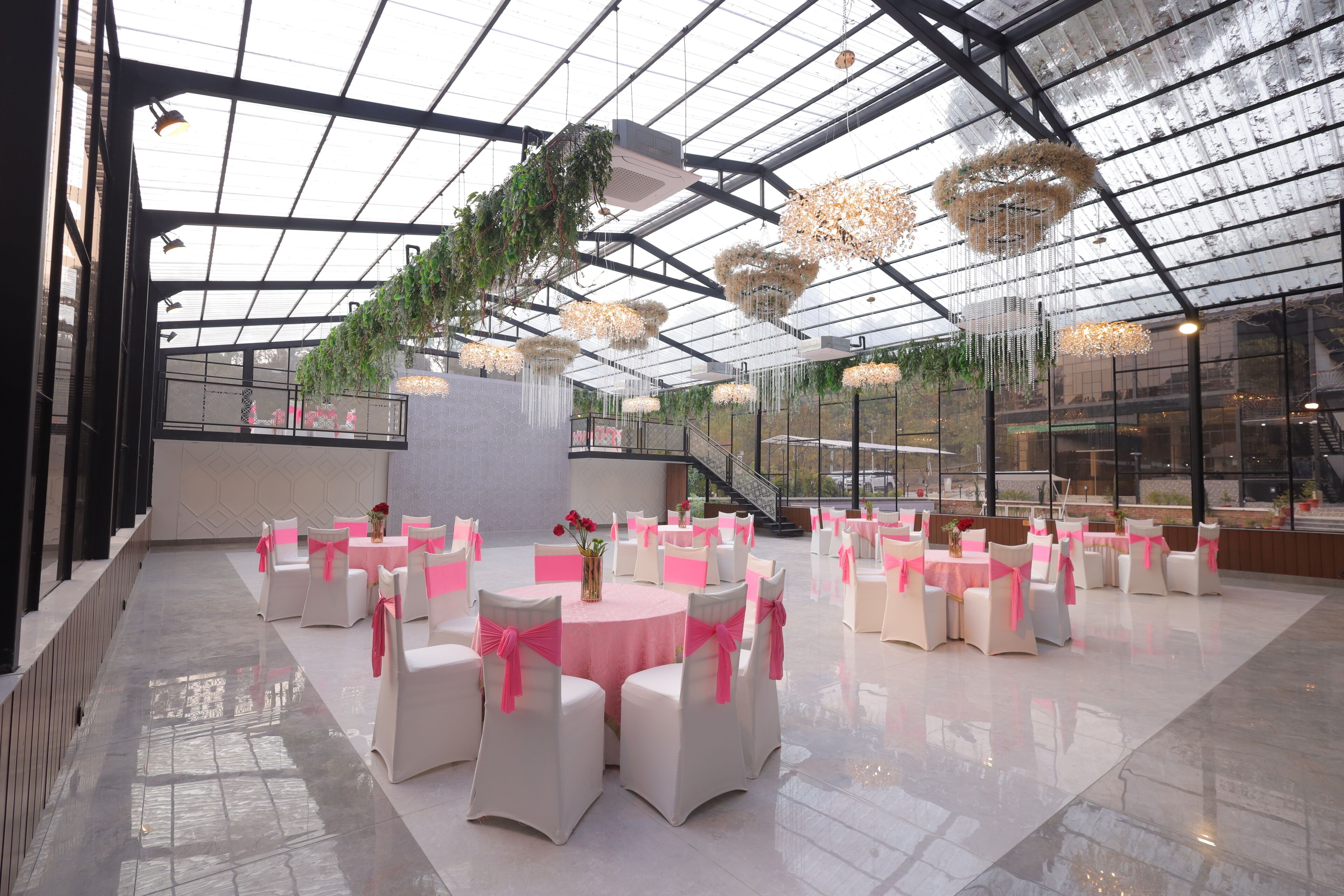 A spacious indoor venue with a high ceiling and glass walls, featuring multiple round tables with chairs, each adorned with pink sashes.