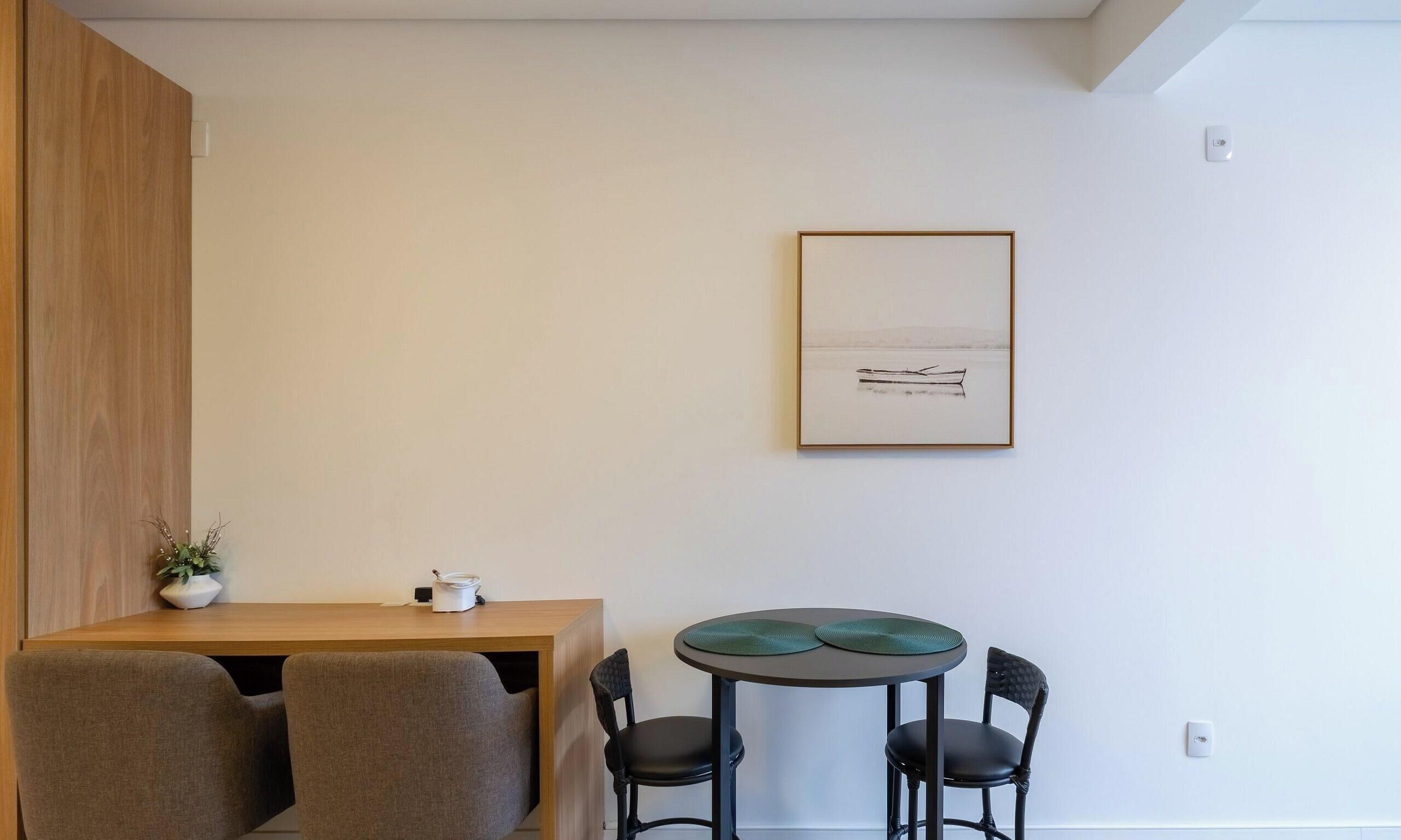Ein schlicht eingerichteter Raum mit einem Holztisch, zwei Stühlen, einem kleinen Tisch mit einer grünen Unterlage und einem gerahmten Bild an der Wand.