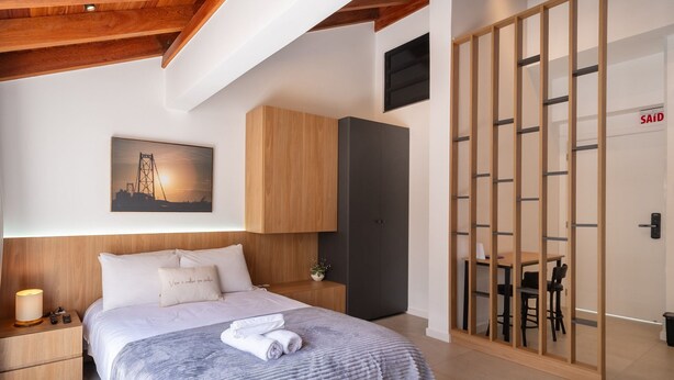 A modern bedroom with a bed, nightstands, a wooden headboard, a painting, and a sliding door with a table and chairs. - Complete Studios in Downtown Floripa - CMA (Florianópolis)