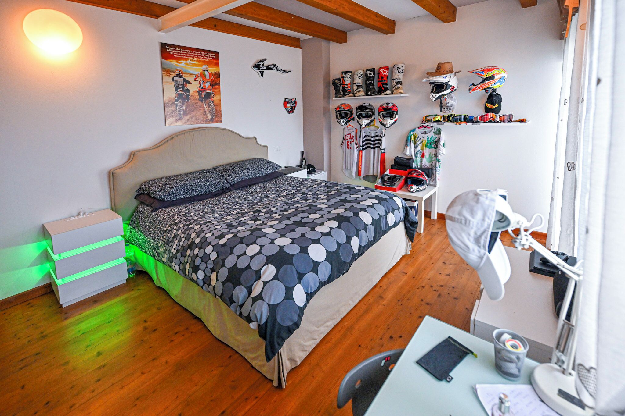 Un dormitorio con una cama, un escritorio, una silla, una lámpara y una estantería con cascos.