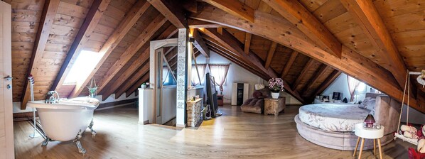 A high-ceilinged attic room with exposed wooden beams, a white bathtub, a round bed, and a small table with a lamp. - Host Monti del Sole (Santa Giustina)