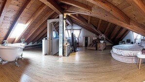 Ein Dachzimmer mit Holzbalken, einer Badewanne, einem runden Bett und einem Essbereich.