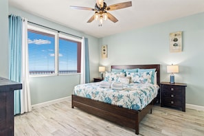 A bedroom with a wooden bed, two bedside tables, a ceiling fan, and a window with blinds. - Shores of Panama 2212 Sea You Soon (Panama City Beach)