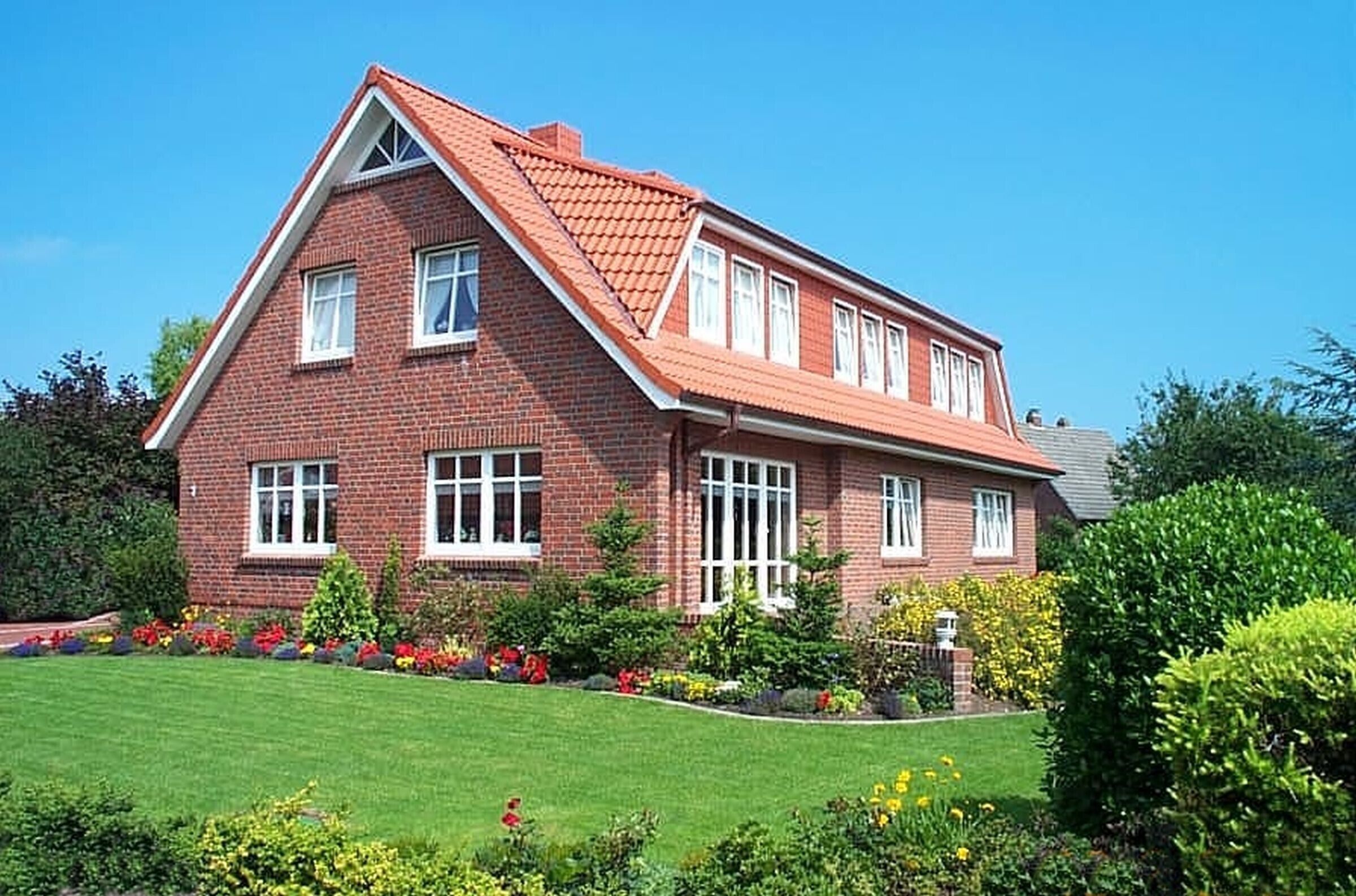 Une maison à deux étages, construite en briques, avec un toit rouge, des fenêtres blanches et un jardin bien entretenu où poussent diverses plantes et fleurs.