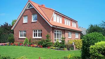 Une maison à deux étages, construite en briques, avec un toit rouge, des fenêtres blanches et un jardin bien entretenu où poussent diverses plantes et fleurs.