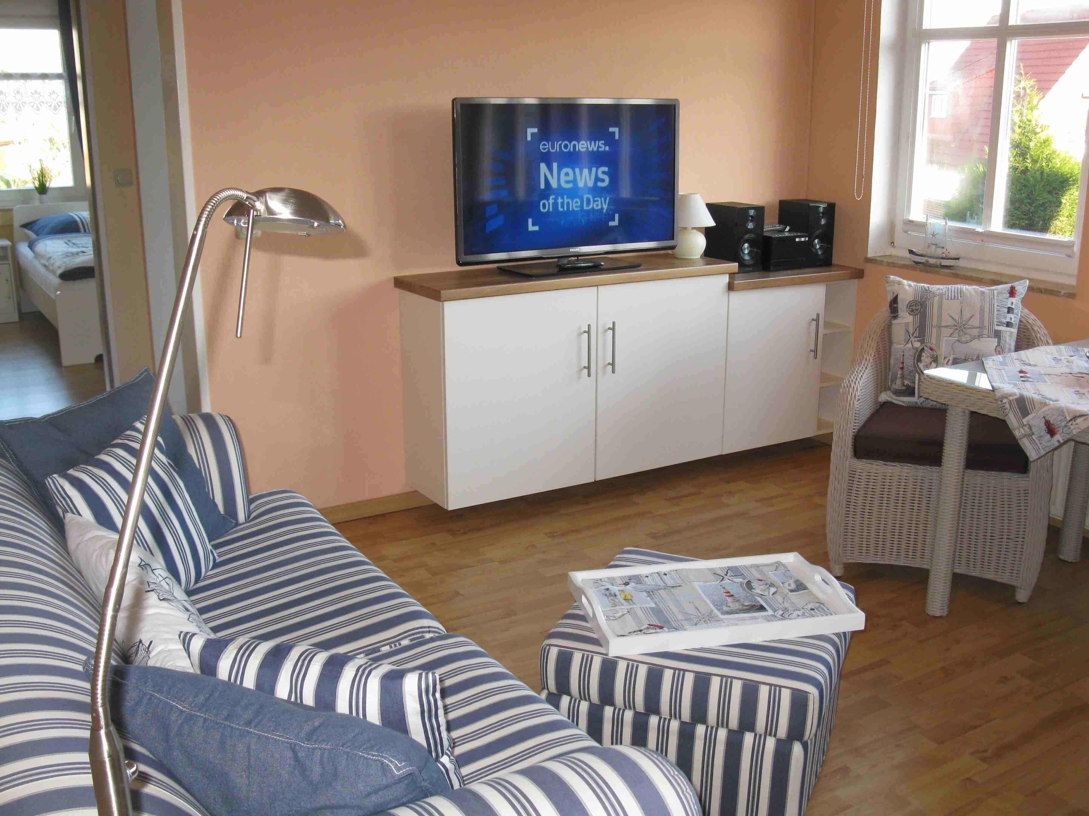 Un salon avec un canapé à rayures, un téléviseur posé sur un meuble, une lampe et un fauteuil en osier.