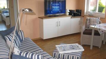 Un salon avec un canapé à rayures, un téléviseur posé sur un meuble, une lampe et un fauteuil en osier.