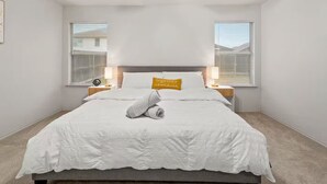 Een opgemaakt bed met een witte dekbedovertrek en een opgerolde handdoek, twee nachtkastjes met lampen, en een plafondventilator.