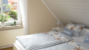 Una camera da letto con soffitto a falda, una finestra vista esterna, un comodino con lampada e un letto con lenzuola a righe e cuscini a forma di stella marina.