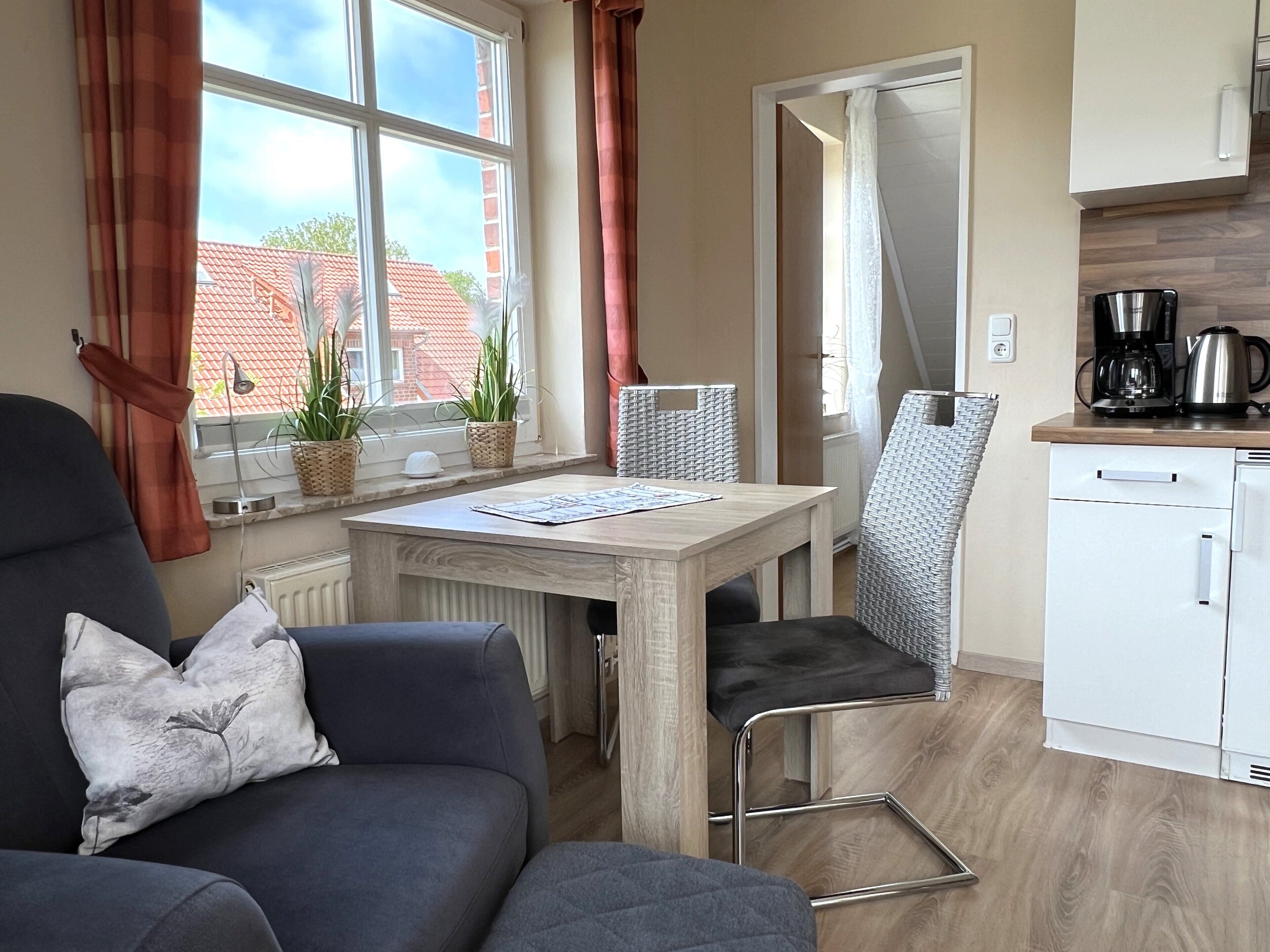 A compact living space with a sofa, a small dining table, a kitchen area with a coffee maker, and a view of a red-tiled building.