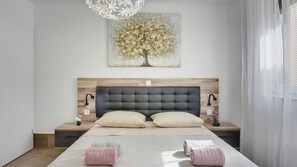 Ein modernes Schlafzimmer mit einem hölzernen Kopfteil, zwei Nachttischlampen, einem großen Gemälde mit einem Baum und einem Bett mit zwei Kissen und zwei zusammengerollten Handtüchern.