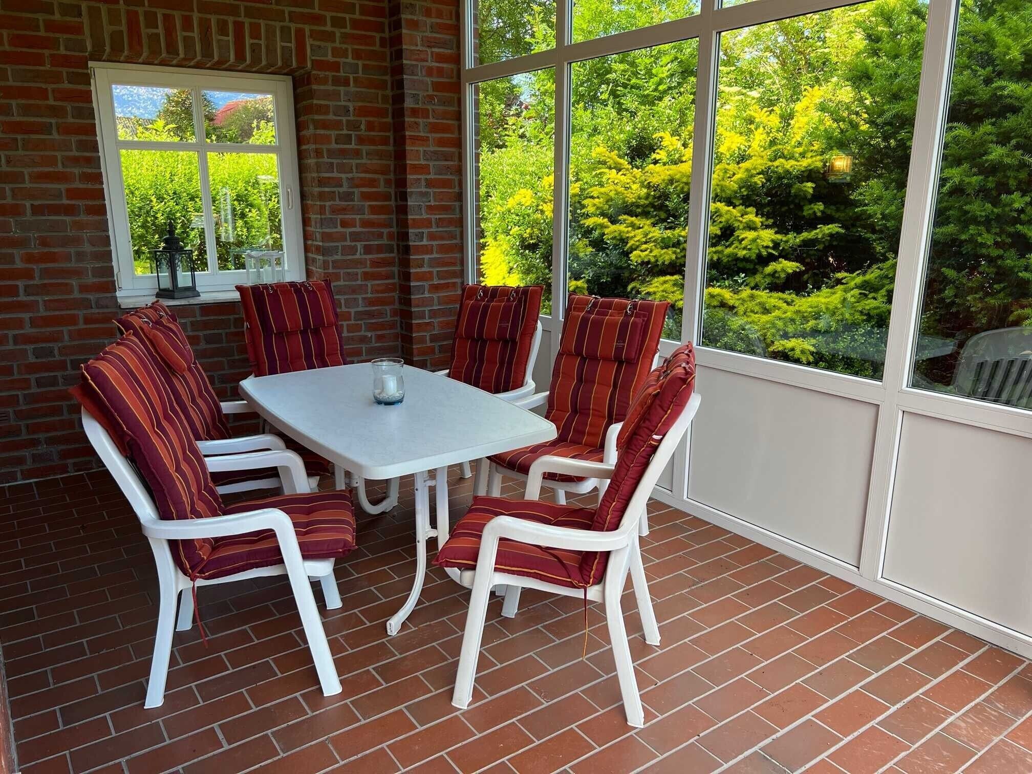 Eine Terrasse mit weißem Tisch und Stühlen, Backsteinboden und großen Fenstern mit Blick auf Grünflächen.