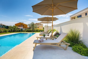 Une piscine entourée de verdure et de bâtiments, avec des chaises longues et des parasols.
