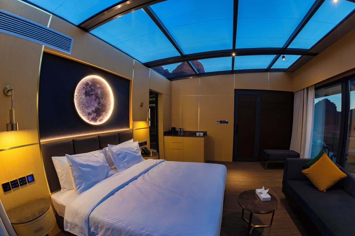 Une chambre d’hôtel moderne dotée d’un grand lit, d’un canapé, d’une petite table, d’une salle de bain et offrant une vue sur les montagnes grâce à une lucarne.