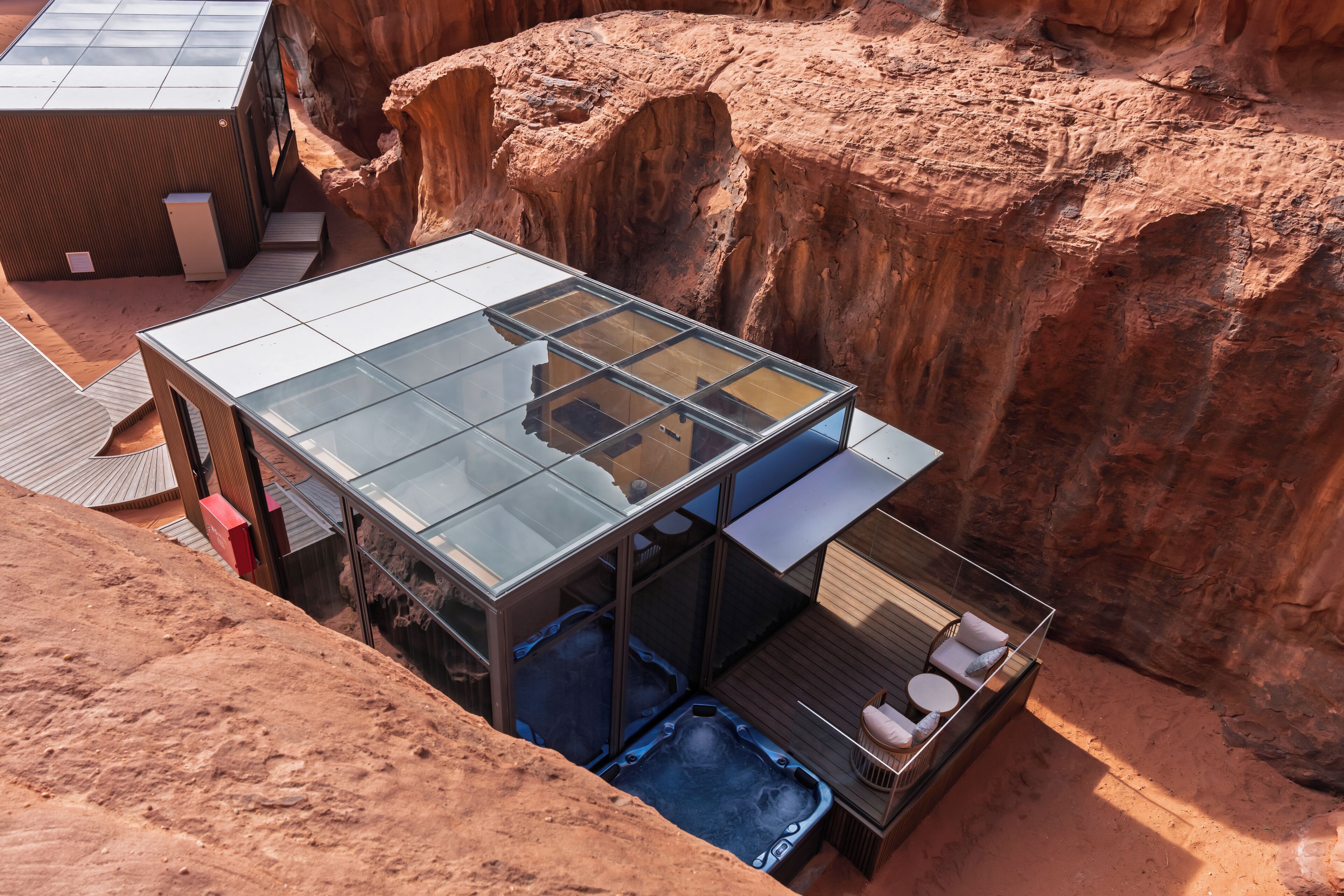 A glass-enclosed modern building with a hot tub and outdoor seating area, set against a rocky canyon backdrop.